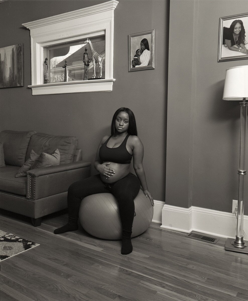 Black and white photograph by Deana Lawson of a pregnant woman sitting on a medicine ball in living room