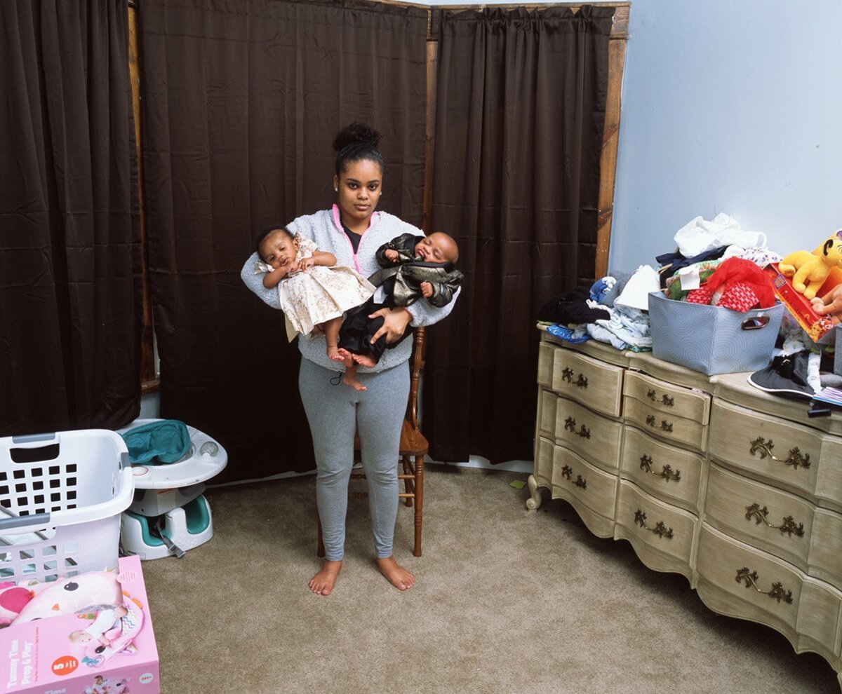 Color photograph by Deana Lawson of a woman holding twin infants standing in a bedroom